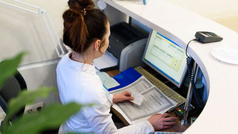 A woman works at a computer with notes and documents in her hand/nearby.